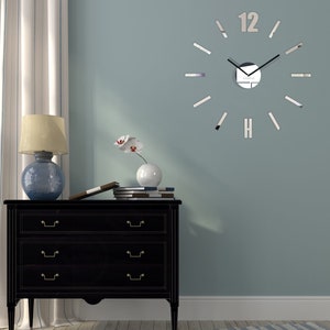 May include: A large, silver, mirrored clock with black hands and Roman numeral markings on a light blue wall. The clock is mounted above a black dresser with three drawers. A white orchid in a white vase sits on top of the dresser.