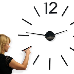 May include: A modern wall clock with black numerals and hour markers. The clock's hands are black, and the clock is being installed on a white wall. A woman is pointing at the clock.