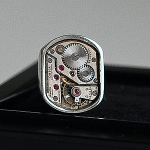 Op de afbeelding: Een zilveren ring met een horloge-mechanisme ontwerp. De ring heeft een zichtbaar horloge-mechanisme met tandwielen en robijnen.