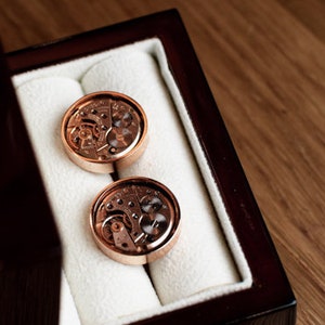 May include: Two gold cufflinks with intricate clockwork designs displayed in a dark wooden box with a white velvet lining.