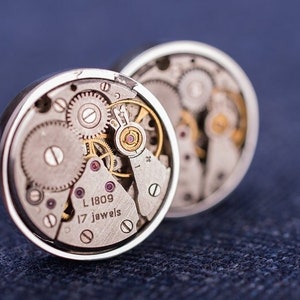 Peut inclure: Une paire de boutons de manchette en argent avec des mécanismes de montre apparents. Les boutons de manchette présentent un boîtier en argent avec une fenêtre transparente révélant le fonctionnement interne d'un mouvement de montre. Le texte "L 1809 17 jewels" est visible sur la face des boutons de manchette.