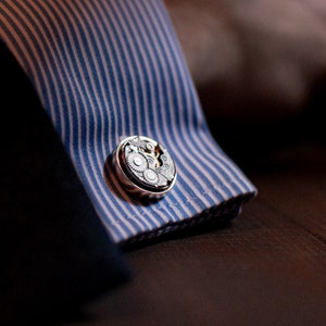 May include: A pair of silver cufflinks with a detailed gear design. The cufflinks are attached to a blue and white striped shirt cuff.