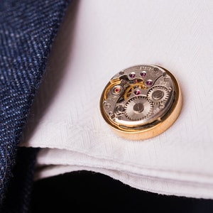 May include: A gold cufflink with a silver watch mechanism inside. The cufflink is on a white fabric surface.
