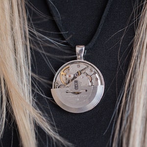Puede incluir: Un collar de plata con un colgante redondo que presenta un mecanismo de relojería visible.