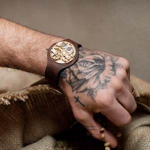 May include: A brown leather wristband with a circular gold-toned metal piece featuring intricate clockwork details. The wristband is worn on a hand with a tattoo.