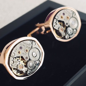 Puede incluir: Un par de gemelos de color dorado con mecanismos de reloj expuestos. Los gemelos están en una caja negra.