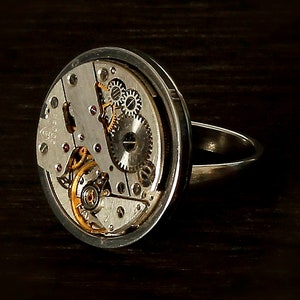 May include: A silver ring with a circular watch face featuring exposed gears and a silver band.