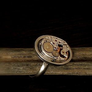 Op de afbeelding: Een zilveren ring met een rond horloge-uurwerk met zichtbare tandwielen en de tekst "Hamilton 17 Jewels 15 Jewels Elec."