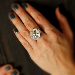 May include: A silver ring with a circular design featuring a watch mechanism. The ring is worn on a person's finger.