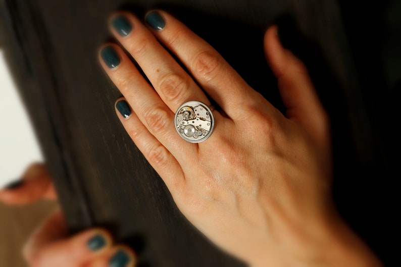 May include: A silver ring with a circular design featuring a watch mechanism. The ring is worn on a person's finger.