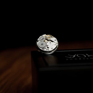 May include: A silver watch movement with exposed gears and the text "Swiss Made" on the face. The movement is sitting on a black box with the word "WOLF" printed on it.