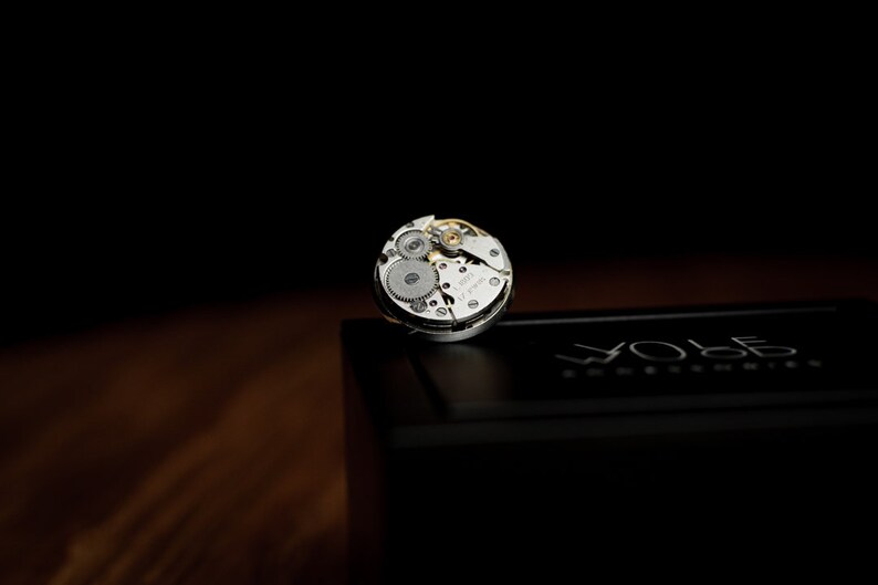 May include: A silver watch movement with exposed gears and the text "Swiss Made" on the face. The movement is sitting on a black box with the word "WOLF" printed on it.