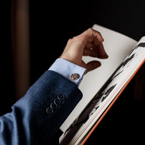 May include: A close-up of a person's wrist wearing a blue suit jacket with a white and blue striped shirt and gold cufflinks. The person's hand is reaching for a book.
