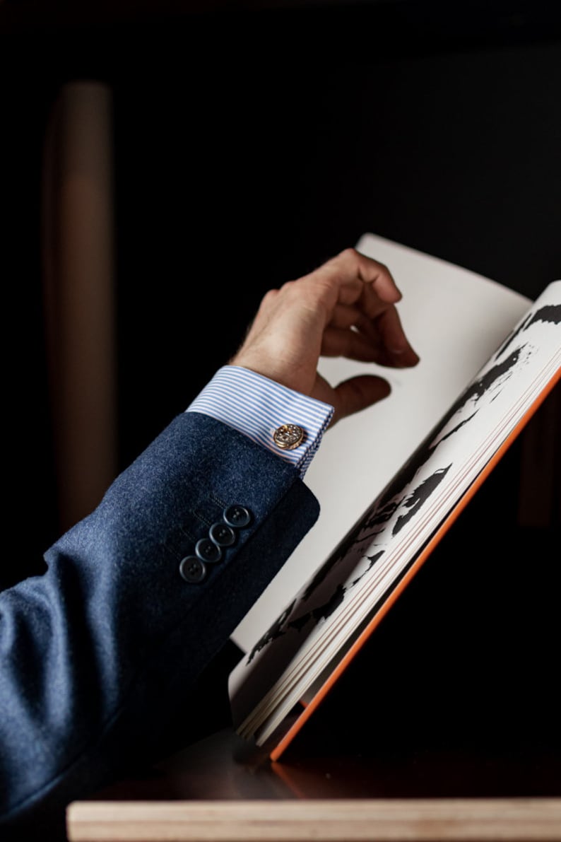 May include: A close-up of a person's wrist wearing a blue suit jacket with a white and blue striped shirt and gold cufflinks. The person's hand is reaching for a book.