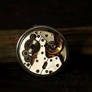 May include: Close-up of a silver watch mechanism with exposed gears and a gold-coloured winding stem. The watch face is not visible.