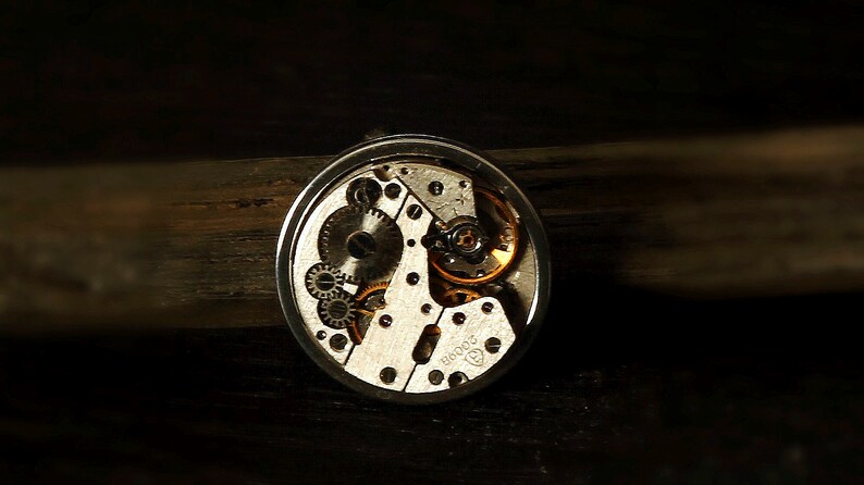 May include: Close-up of a silver watch mechanism with exposed gears and a gold-coloured winding stem. The watch face is not visible.