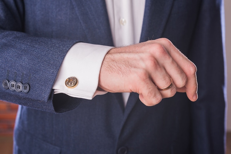 May include: A man wearing a blue suit jacket with a white shirt and cufflinks. He is wearing a gold wedding band on his ring finger.