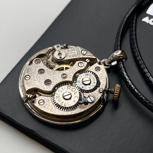 May include: A silver watch movement pendant with detailed gears and a small gold crown. The pendant is on a black cord necklace. The watch mechanism is visible, showcasing the inner workings of the timepiece.