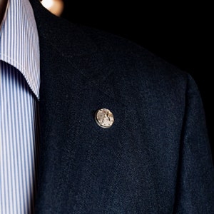 May include: A dark blue blazer with a herringbone pattern and a small silver pin with a clock face design.