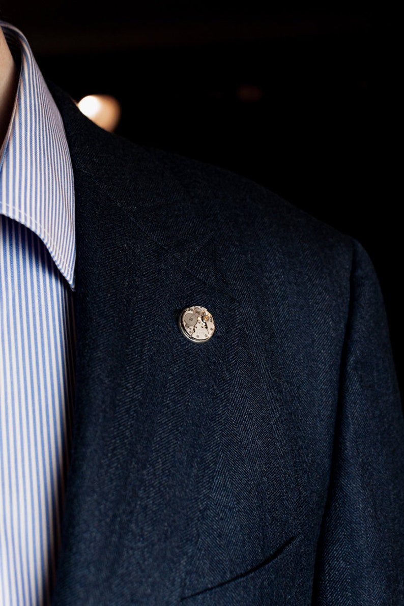 May include: A dark blue blazer with a herringbone pattern and a small silver pin with a clock face design.