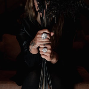 May include: A person wearing a black leather jacket and black trousers is holding a bouquet of black feathers. They are wearing three silver rings with intricate designs on their fingers.