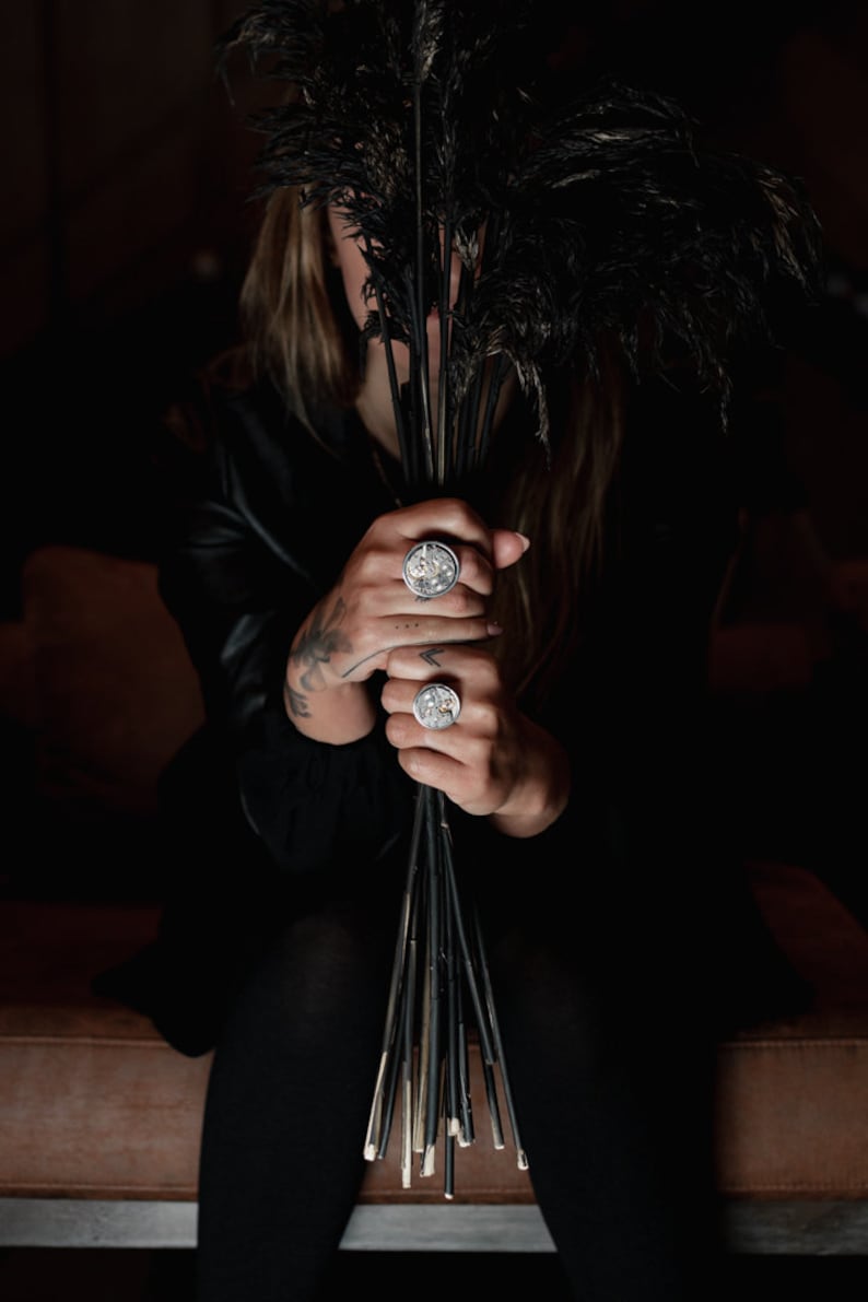 May include: A person wearing a black leather jacket and black trousers is holding a bouquet of black feathers. They are wearing three silver rings with intricate designs on their fingers.