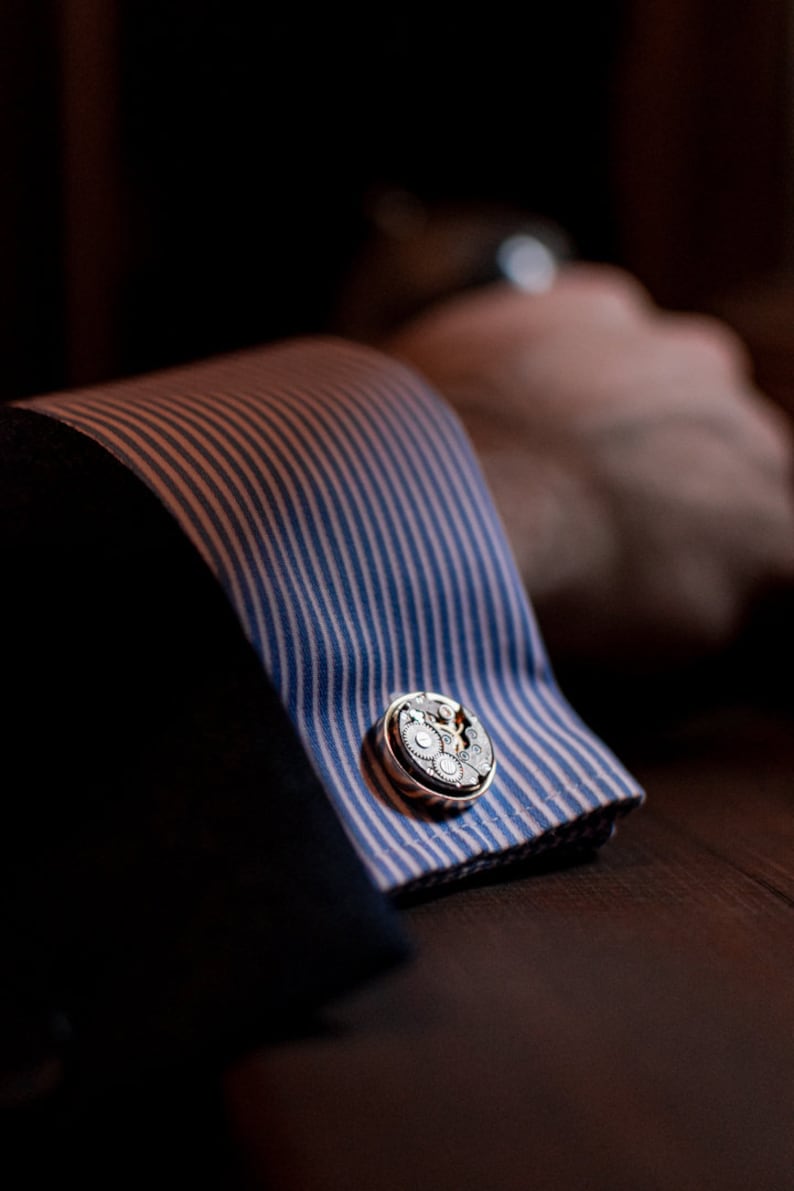 May include: A pair of silver cufflinks with a detailed gear design. The cufflinks are attached to a blue and white striped shirt cuff.