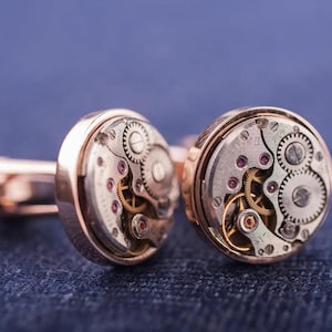May include: A pair of gold cufflinks with a circular design featuring exposed watch gears. The gears are visible through a clear glass or plastic cover.