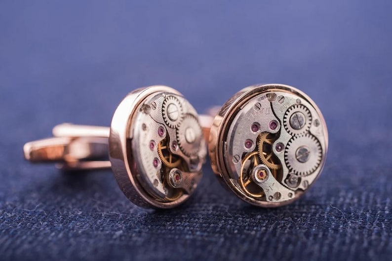 May include: A pair of gold cufflinks with a circular design featuring exposed watch gears. The gears are visible through a clear glass or plastic cover.