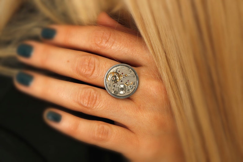 May include: A silver ring featuring a circular watch face design. The watch face is open, revealing the inner workings of the watch.
