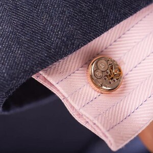 May include: Gold cufflinks with a detailed clockwork design. The cufflinks are on a pink and white striped shirt cuff.