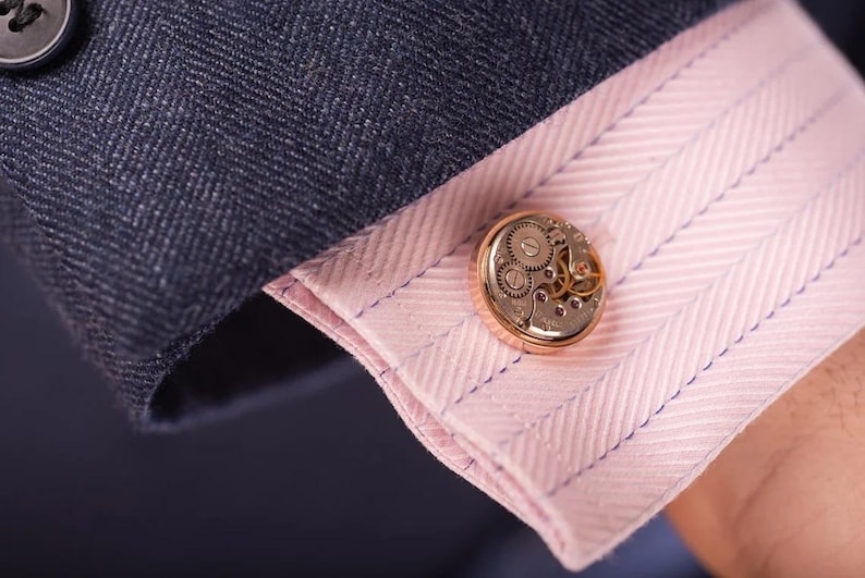 May include: Gold cufflinks with a detailed clockwork design. The cufflinks are on a pink and white striped shirt cuff.