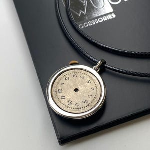 May include: A silver pendant necklace with a vintage watch face, black numerals on a beige face, and a black cord. The pendant is displayed on a black surface with the brand name "WOLF WOOD ACCESSORIES".