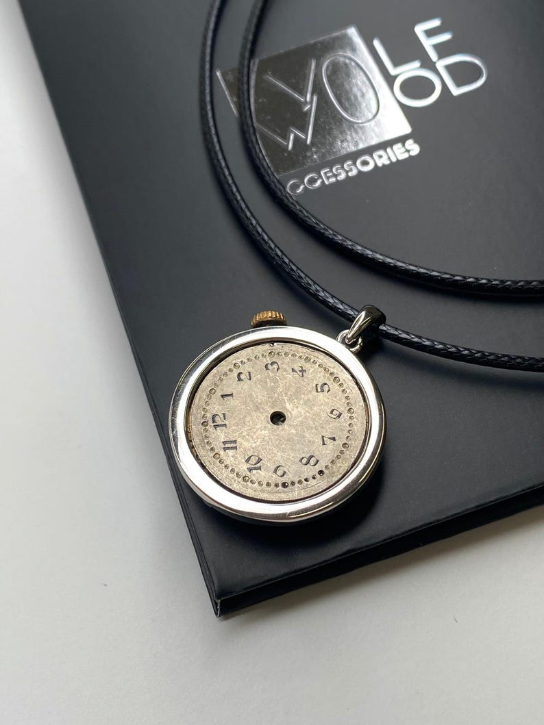 May include: A silver pendant necklace with a vintage watch face, black numerals on a beige face, and a black cord. The pendant is displayed on a black surface with the brand name "WOLF WOOD ACCESSORIES".