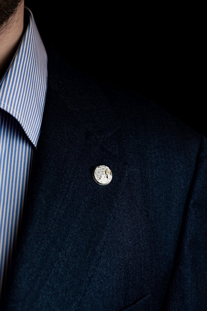 May include: A dark blue blazer with a small, round, silver pin featuring a detailed clockwork design.