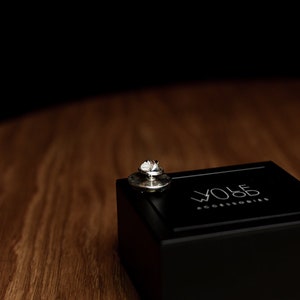 May include: A silver pin with a small, round design sits on top of a black box. The box has the text "WOLF ACCESSORIES" printed on the top.