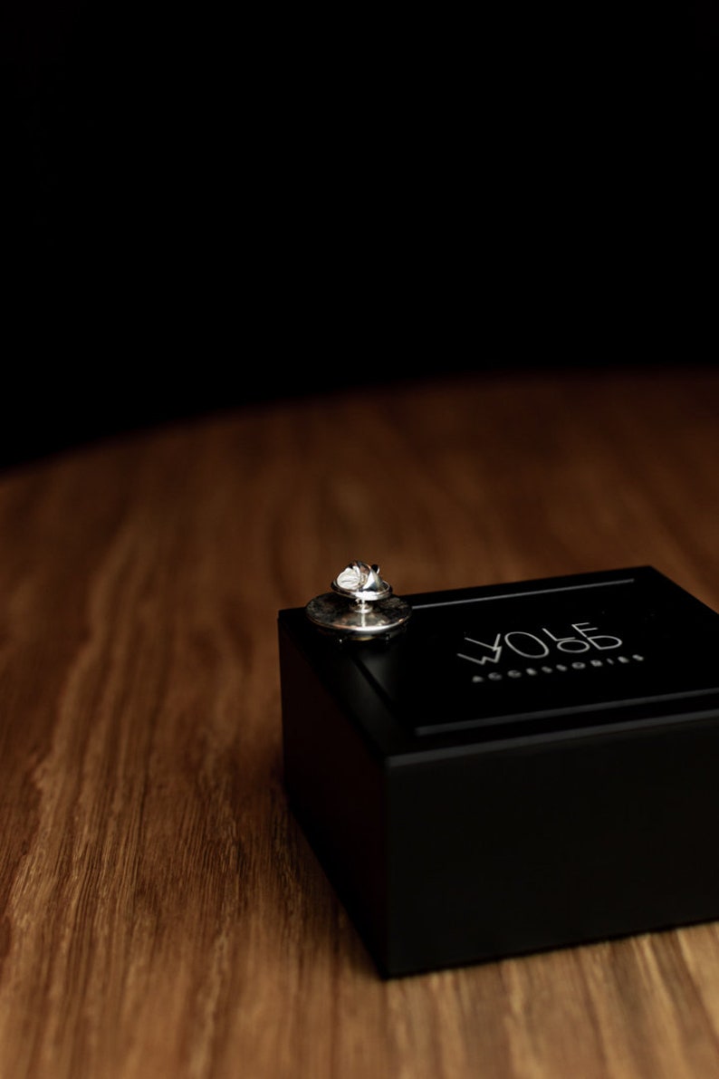 May include: A silver pin with a small, round design sits on top of a black box. The box has the text "WOLF ACCESSORIES" printed on the top.