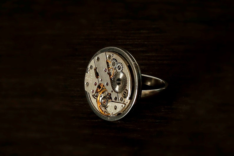 May include: A silver ring with a circular watch face featuring exposed gears and a silver band.