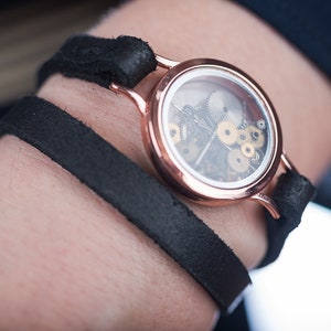 May include: A rose gold watch with a clear face revealing the inner workings of the watch mechanism. The watch is attached to a black leather band.