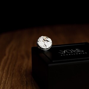 May include: A silver watch movement on a black box with the text "WOLF  watch winders" on the top.