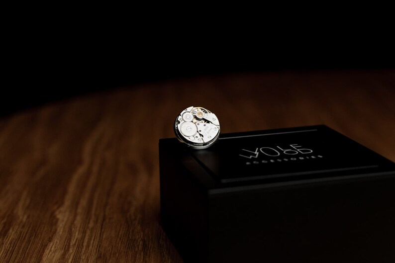 May include: A silver watch movement on a black box with the text "WOLF  watch winders" on the top.