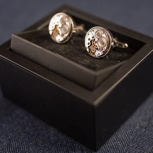 May include: A pair of silver cufflinks with a black box. The cufflinks have a circular design with visible gears and a silver finish.