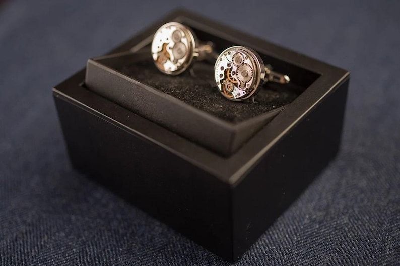May include: A pair of silver cufflinks with a black box. The cufflinks have a circular design with visible gears and a silver finish.