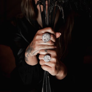May include: Two silver rings featuring intricate clockwork designs. The rings are worn on the fingers of a person holding a bunch of black feathers.