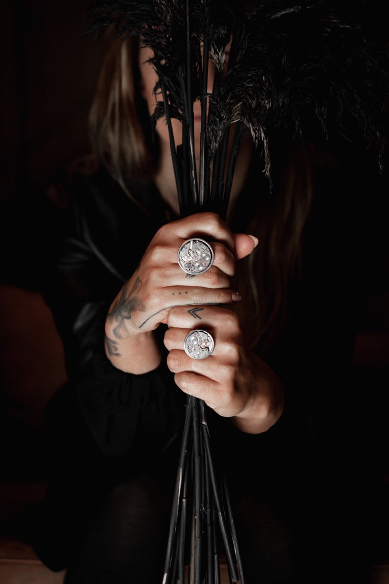 May include: Two silver rings featuring intricate clockwork designs. The rings are worn on the fingers of a person holding a bunch of black feathers.