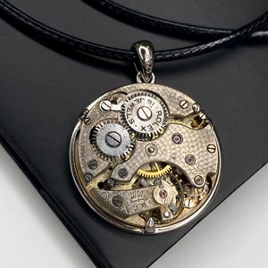 May include: A silver pendant necklace showcasing the internal mechanism of a watch. The circular pendant reveals gears, jewels, and the text "15 JEWELS" and "ROLEX". The pendant is suspended from a black cord.