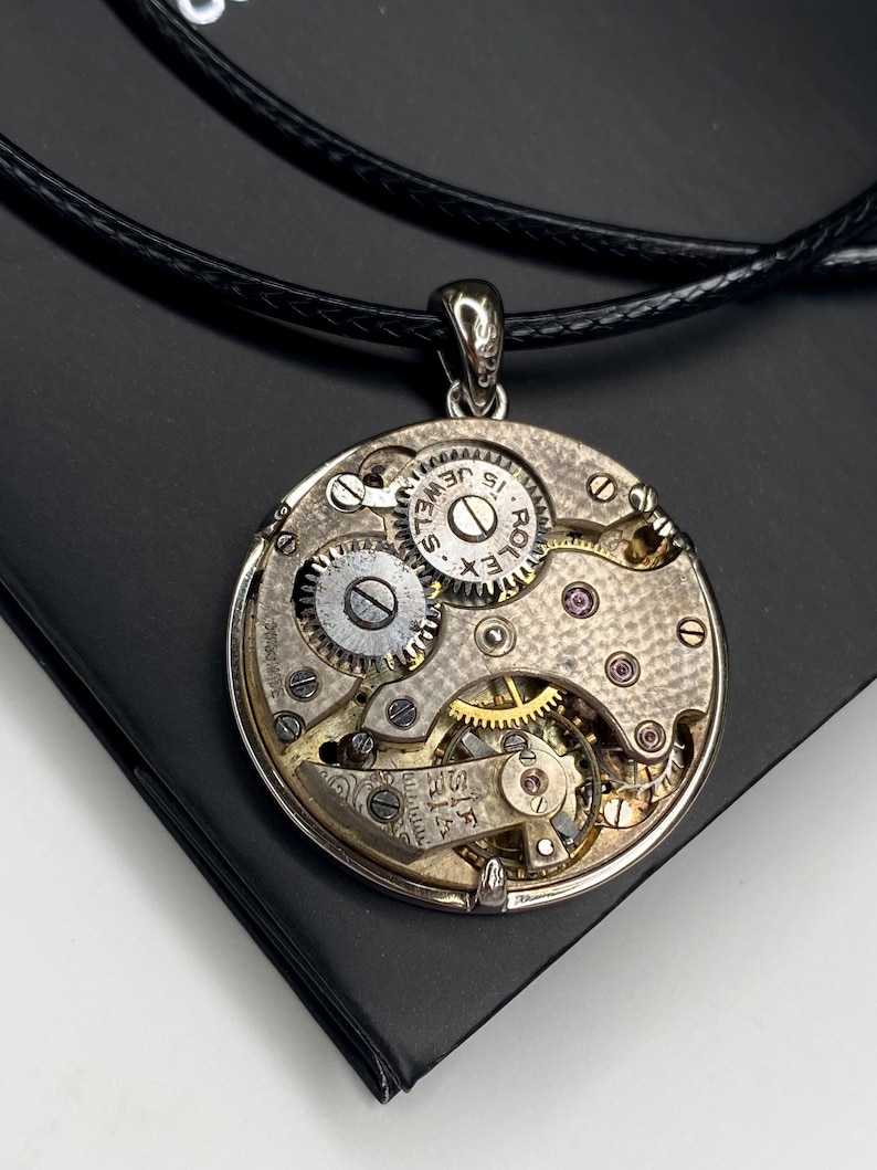 May include: A silver pendant necklace showcasing the internal mechanism of a watch. The circular pendant reveals gears, jewels, and the text "15 JEWELS" and "ROLEX". The pendant is suspended from a black cord.