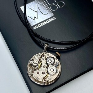 May include: A silver pendant necklace with a repurposed watch mechanism. The round pendant displays intricate gears, springs, and jewels. The necklace has a black leather cord and is presented on a black box with the text "WOLF WOOD ACCESSORIES".