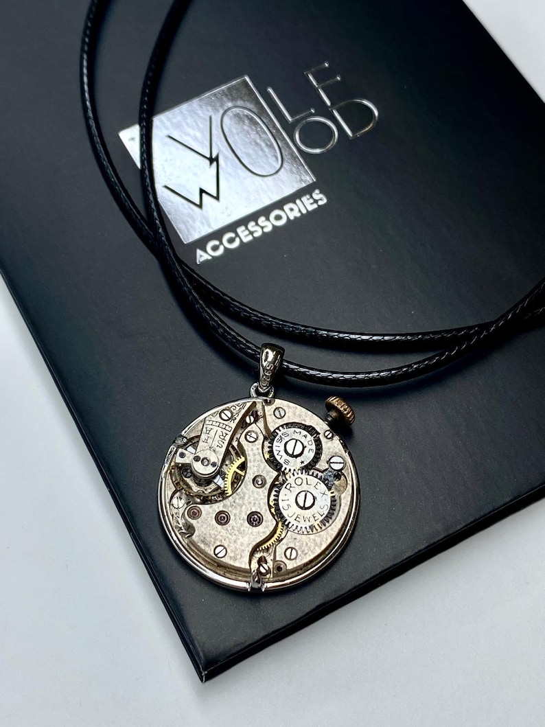 May include: A silver pendant necklace with a repurposed watch mechanism. The round pendant displays intricate gears, springs, and jewels. The necklace has a black leather cord and is presented on a black box with the text "WOLF WOOD ACCESSORIES".