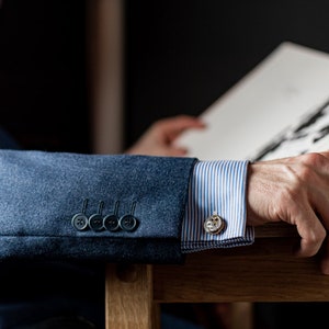 May include: A close-up of a blue blazer sleeve with four black buttons and a white and blue striped shirt cuff with a silver cufflink. The cufflink has a detailed design.
